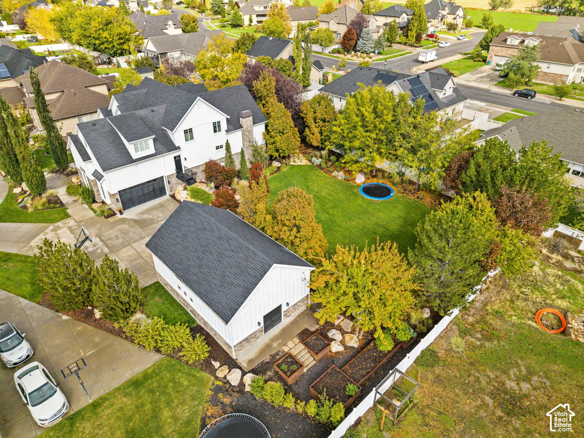 Aerial perspective of a property featuring a white board-and-batten exterior, a detached garage, a concrete driveway, multi-level raised planter beds, and manicured landscaping