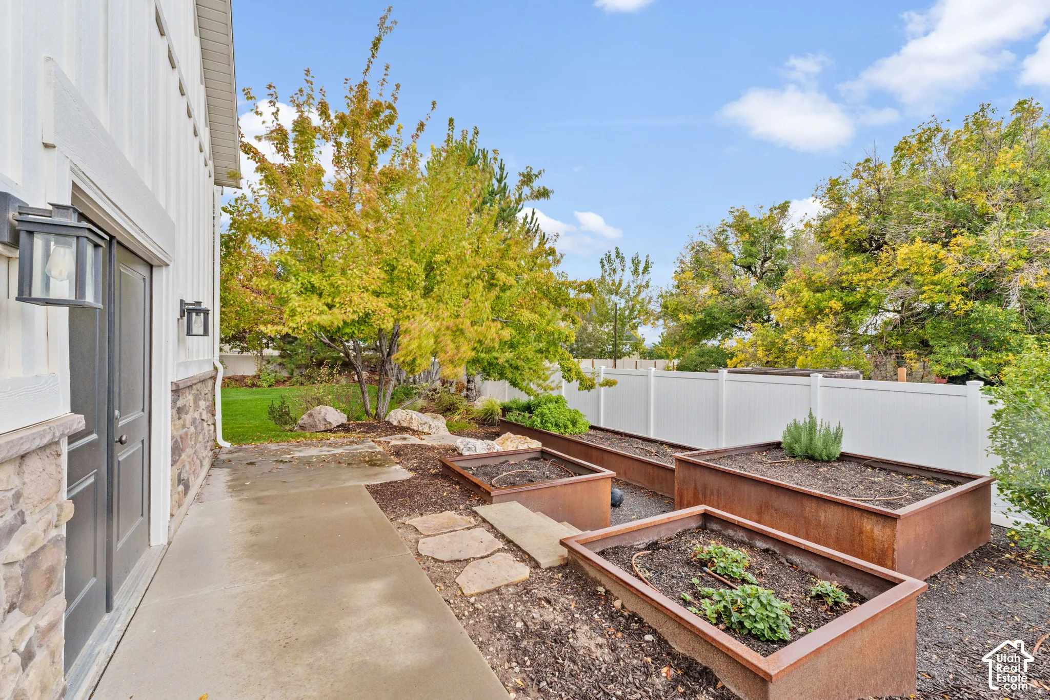 Exterior side yard featuring a concrete pathway, raised metal garden beds, stone hardscaping, and a white privacy fence
