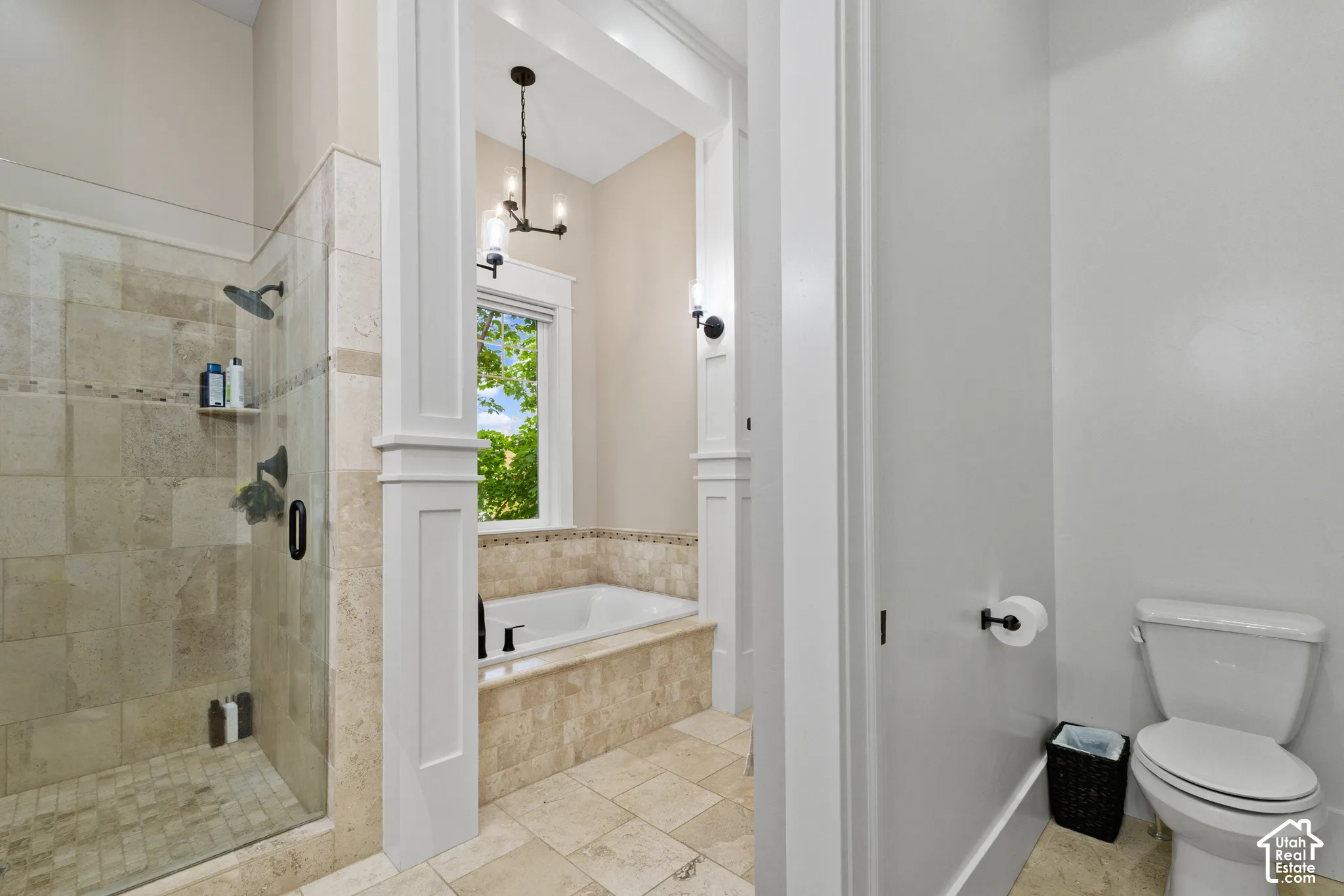 Walk-in shower with stone tile surround, soaking tub with tile deck, decorative millwork columns, and built-in wall lighting fixtures