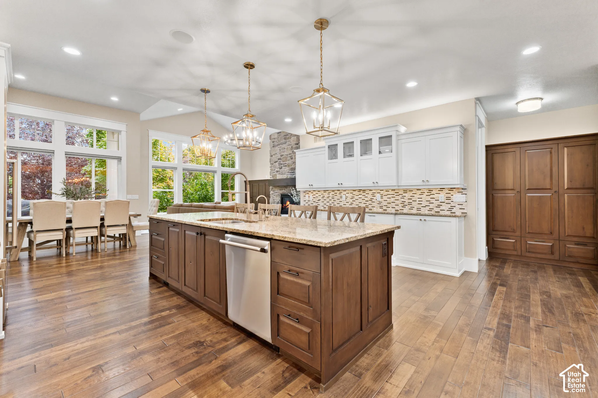 Kitchen featuring wood-finish flooring, a central island with integrated stainless steel dishwasher, stone-tiled backsplash, white upper cabinetry, and decorative metal pendant lighting
