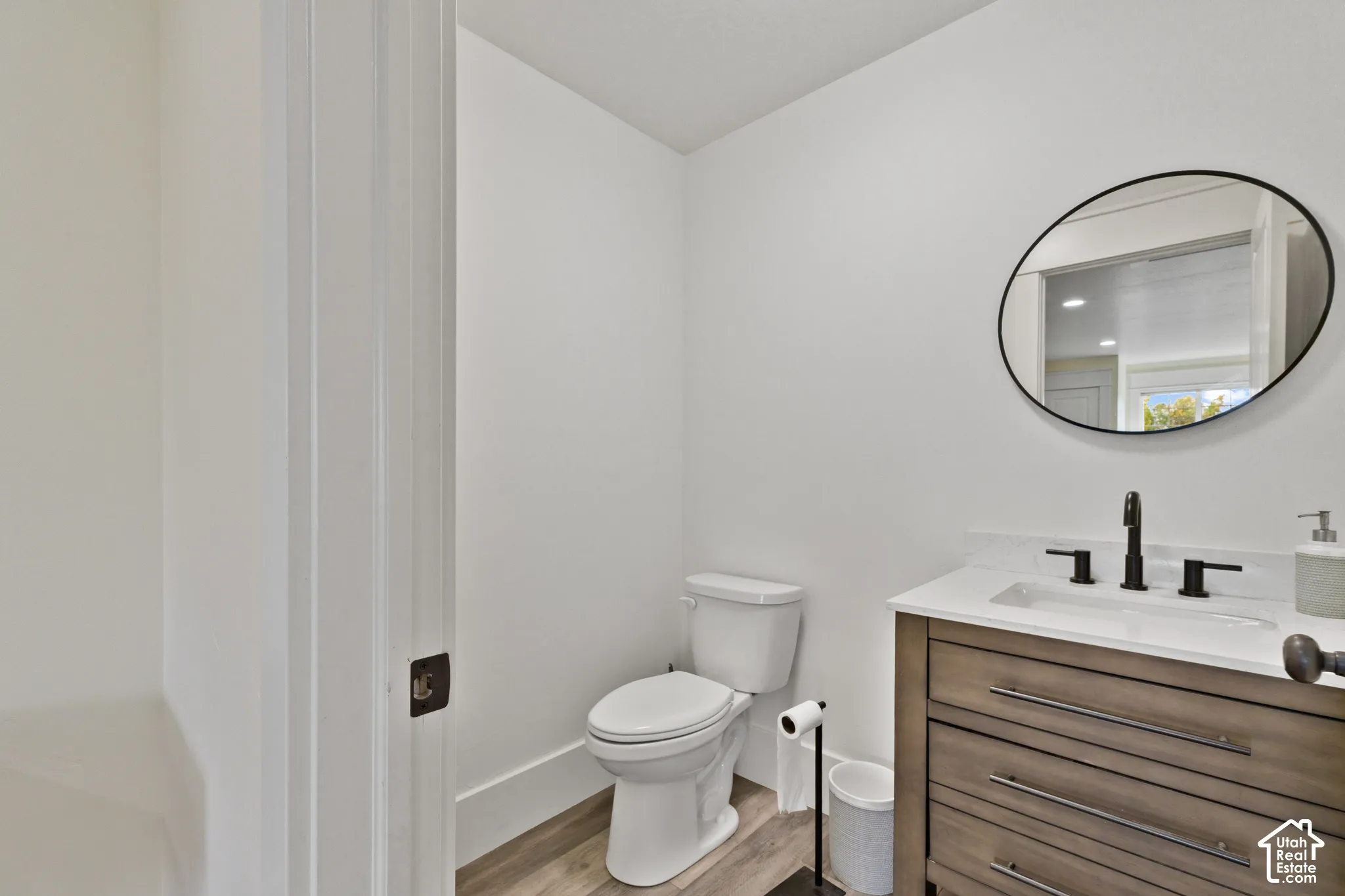 Vanity with wood-finish cabinetry, white countertop, matte black widespread faucet, oval wall-mounted mirror, and wood-finish flooring