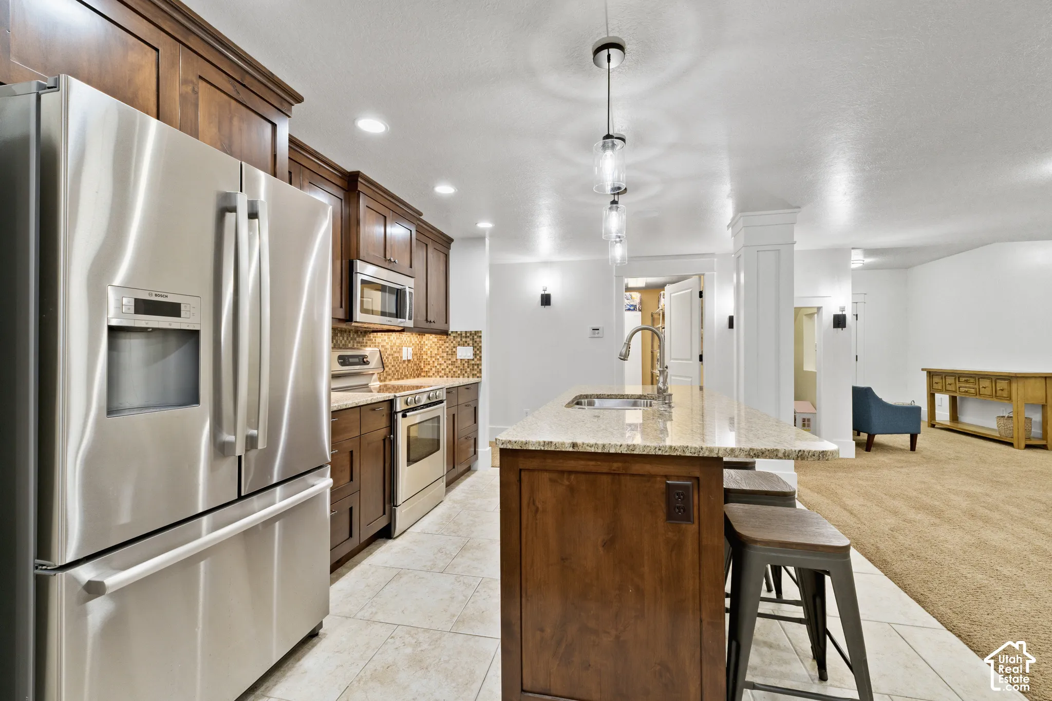 Kitchen featuring dark wood cabinetry, stone-finish countertops, stainless steel appliances, tile flooring, and a central island with an integrated sink and pendant lighting