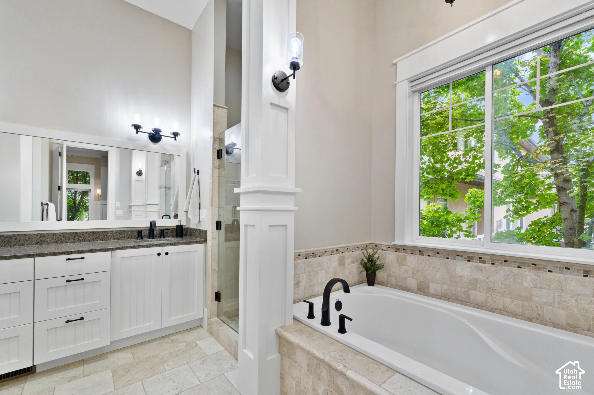 Custom white cabinetry with stone countertops, a soaking tub with stone tile surround, a glass-enclosed shower, wall-mounted lighting fixtures, and a large window