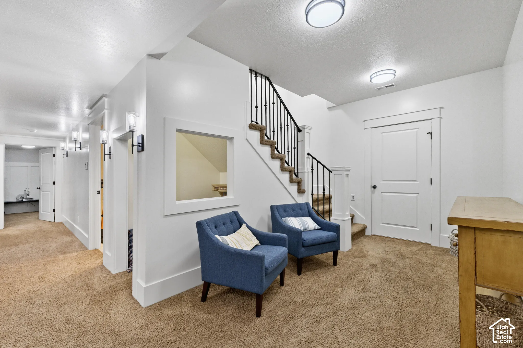 Interior area featuring wall-to-wall carpeting, a built-in square interior window, staircase with wrought iron balusters, multiple wall-mounted sconces, and overhead flush-mount lighting