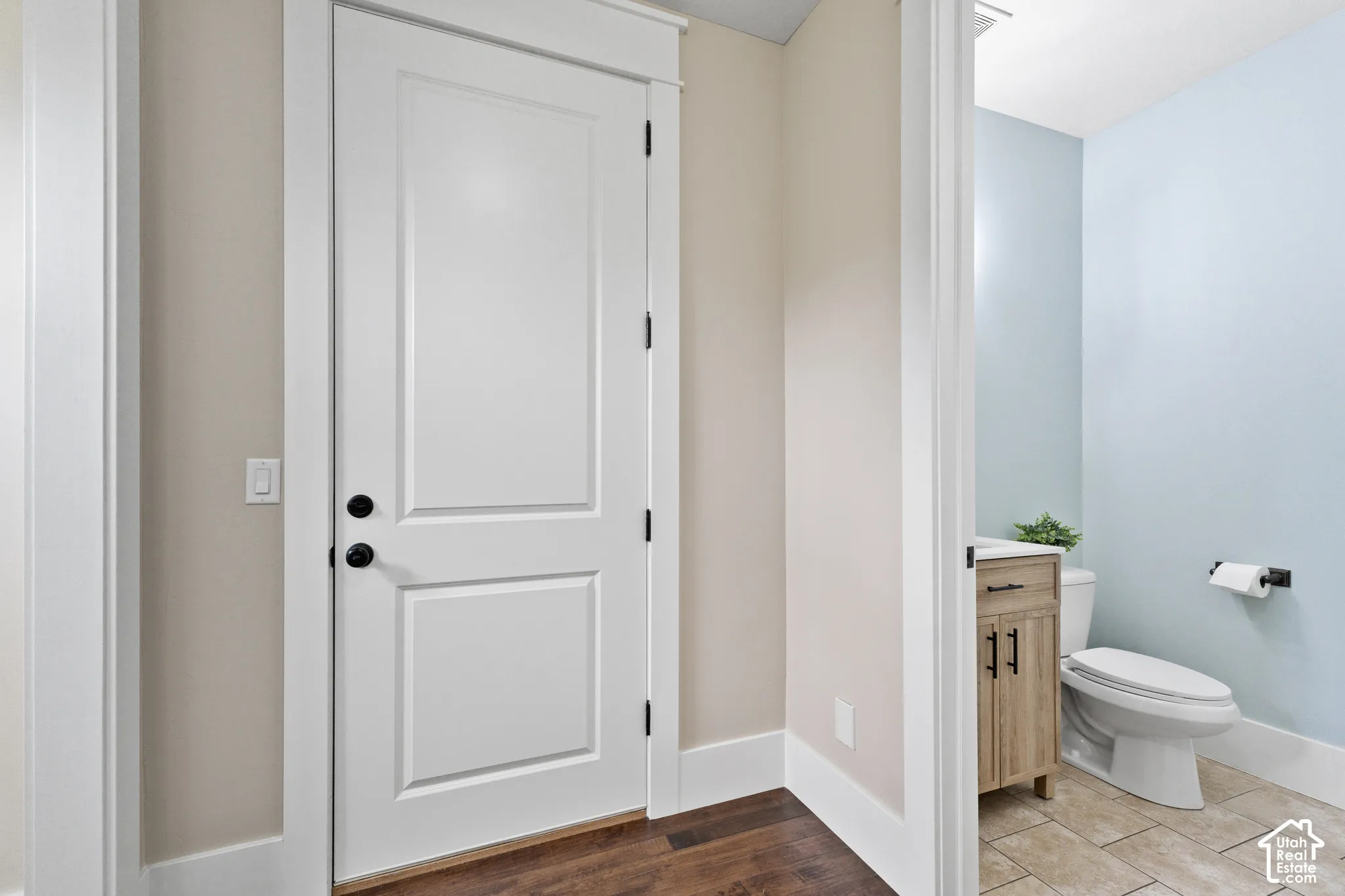 Solid-core door with matte black hardware, wood-finish flooring, wood-tone vanity cabinetry, light-colored tile flooring, and matte black plumbing fixtures
