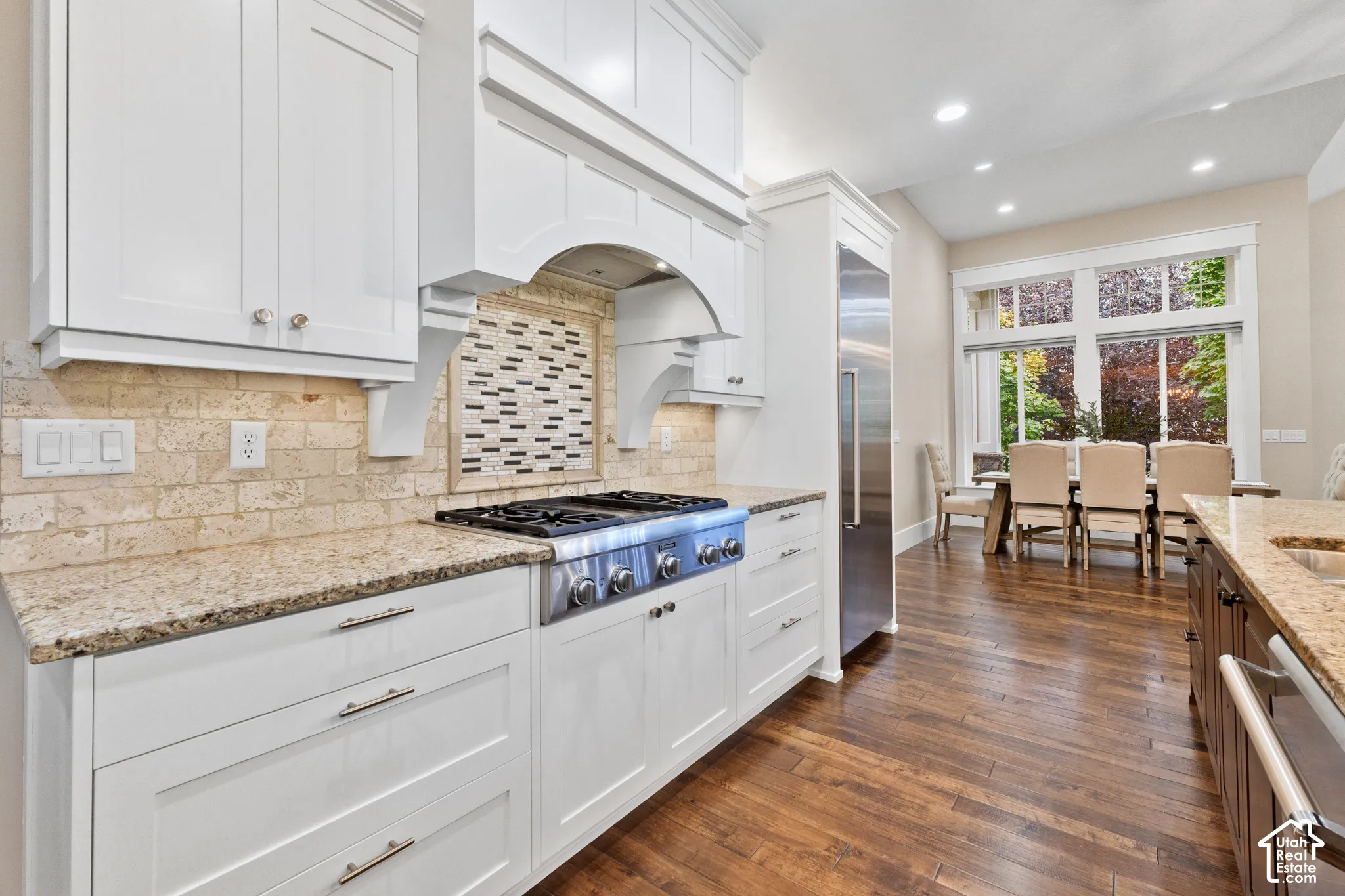 Custom white cabinetry featuring a decorative range hood, stone-finish countertops, tiled backsplash, stainless steel gas range, and wood-finish flooring