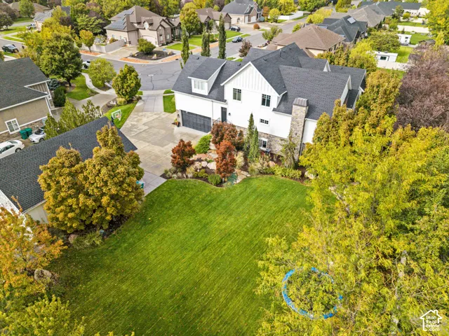 Aerial perspective featuring a multi-story residence with a stone and vertical-siding exterior, expansive grass lawn, mature trees, tiered stone landscaping, and a concrete driveway