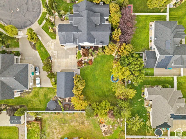 Aerial view of a residential property featuring a large backyard, concrete driveway, multi-gabled roofline, established trees, and perimeter fencing