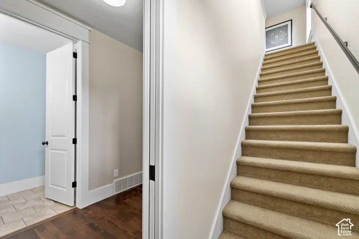 Staircase featuring neutral carpeted treads and a mounted metal handrail, complemented by wood-finish flooring and ceramic tile flooring in the threshold area