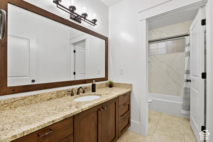 Stained wood cabinetry with stone-finish countertop, undermount sink, three-light fixture, framed vanity mirror, and bathtub enclosure with stone-finish wall surround