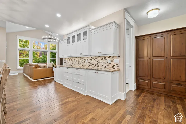 White shaker-style cabinetry with glass-front uppers, stone countertop, mosaic tile backsplash, wood-finish flooring, and floor-to-ceiling dark wood-finish cabinetry