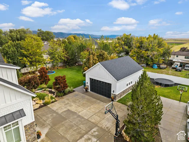 Detached garage with board-and-batten siding and stone masonry accents, expansive concrete motor court, manicured lawn, tiered rock landscaping, and mature foliage
