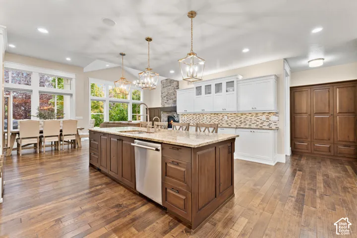 Kitchen featuring wood-finish flooring, a central island with integrated stainless steel dishwasher, stone-tiled backsplash, white upper cabinetry, and decorative metal pendant lighting