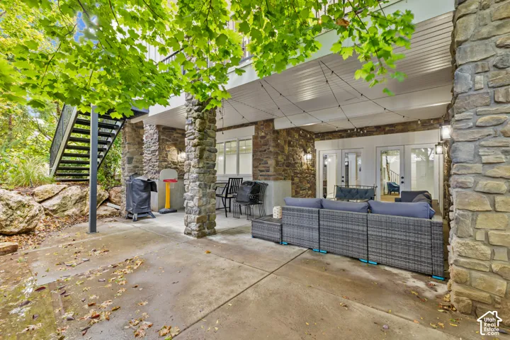 Covered patio featuring stone-clad pillars, concrete flooring, exterior wall-mounted lighting, and a metal staircase