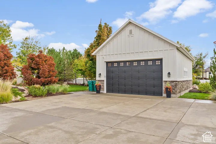 Detached garage featuring white board-and-batten siding, stone veneer accents, an expansive concrete driveway, and a dark-toned multi-panel garage door with transom windows