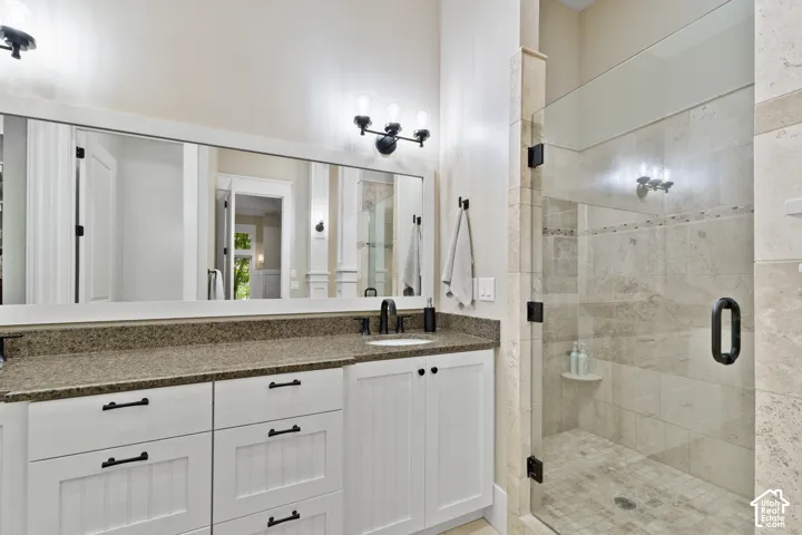 White cabinetry with matte black hardware, stone-finish countertop, frameless glass shower enclosure with stone tile surround, and wall-mounted vanity lighting