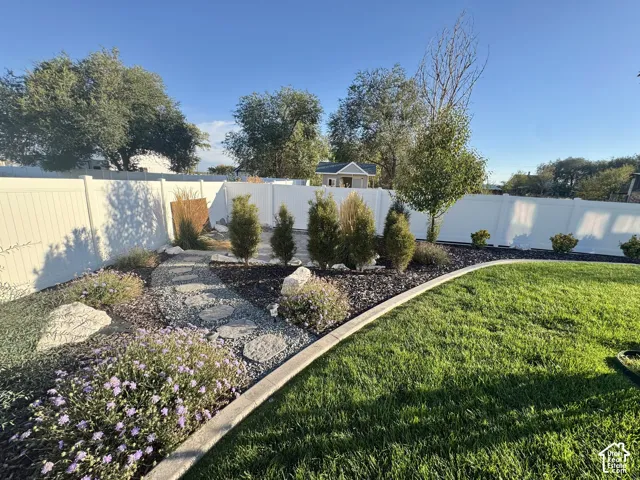 Landscaped backyard featuring a white vinyl privacy fence, a natural stone walking path, mature ornamental shrubbery, decorative rock mulch, and a grass lawn
