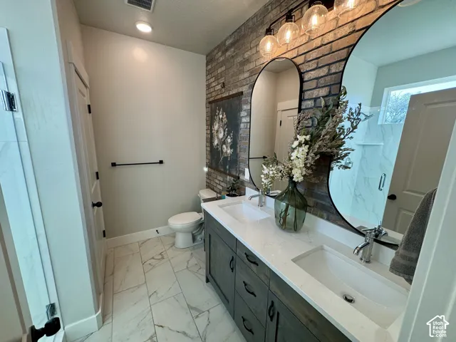 Bathroom featuring a double vanity with quartz countertops, undermount sinks, wood-finish cabinetry, floor-to-ceiling brick accent wall, and oversized arched mirrors