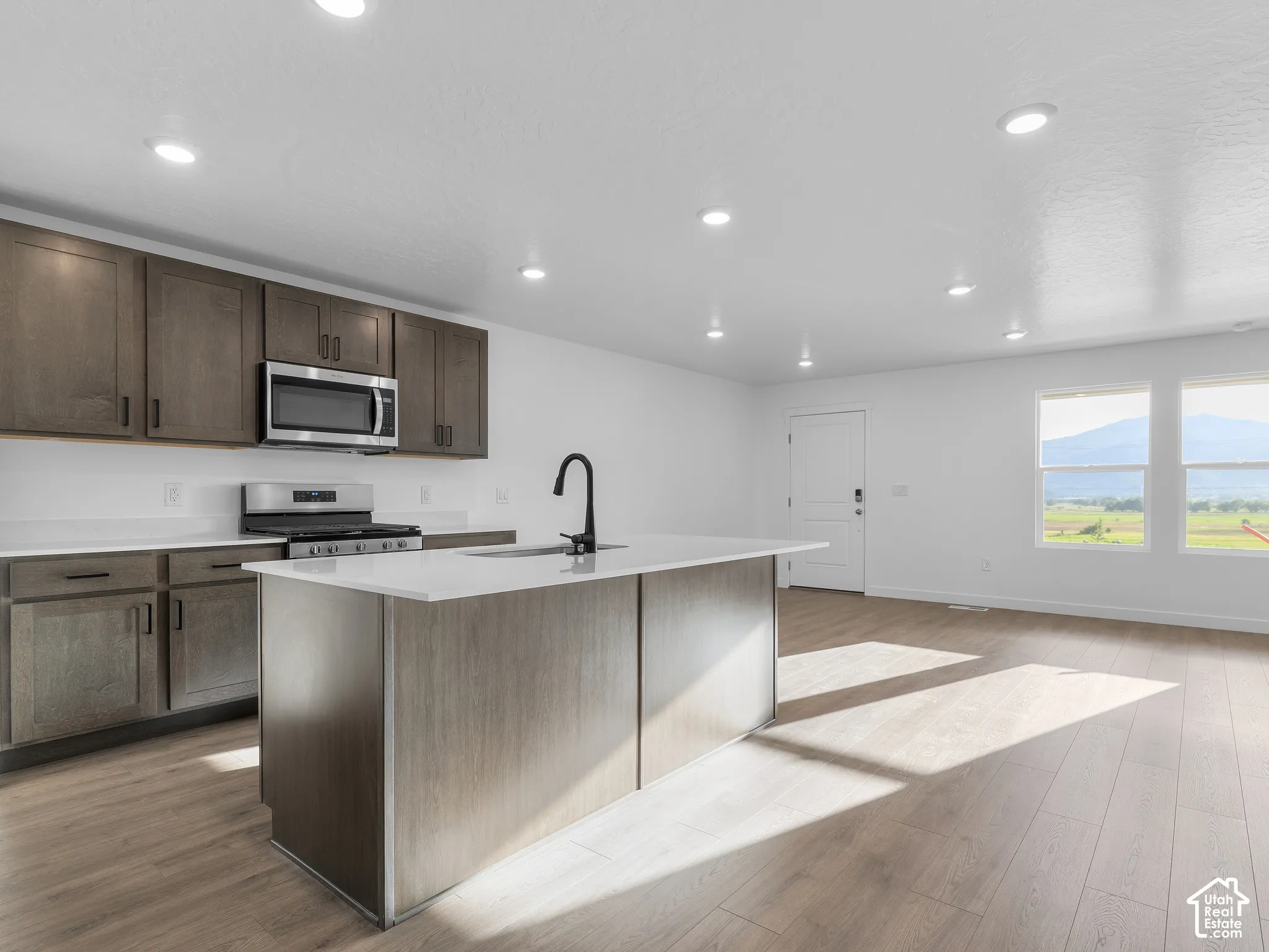 Kitchen featuring dark-toned cabinetry, white stone-composite countertops, a central island with a matte black faucet, recessed lighting, and wood-finish flooring