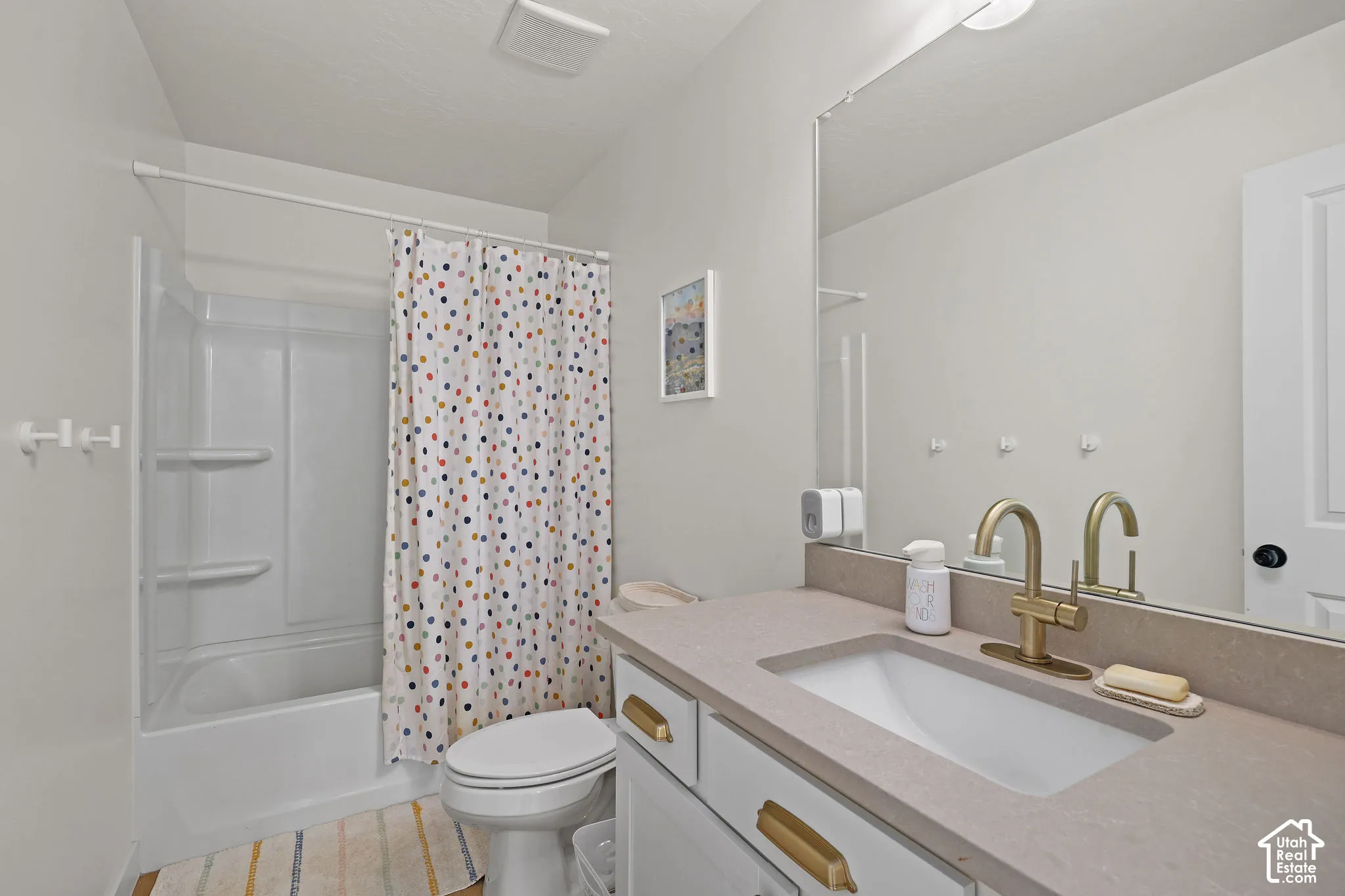 Bathroom featuring a vanity with a neutral-toned solid surface countertop, an undermount sink, and brass-finish hardware, paired with a white tub and shower combination with integrated shelving