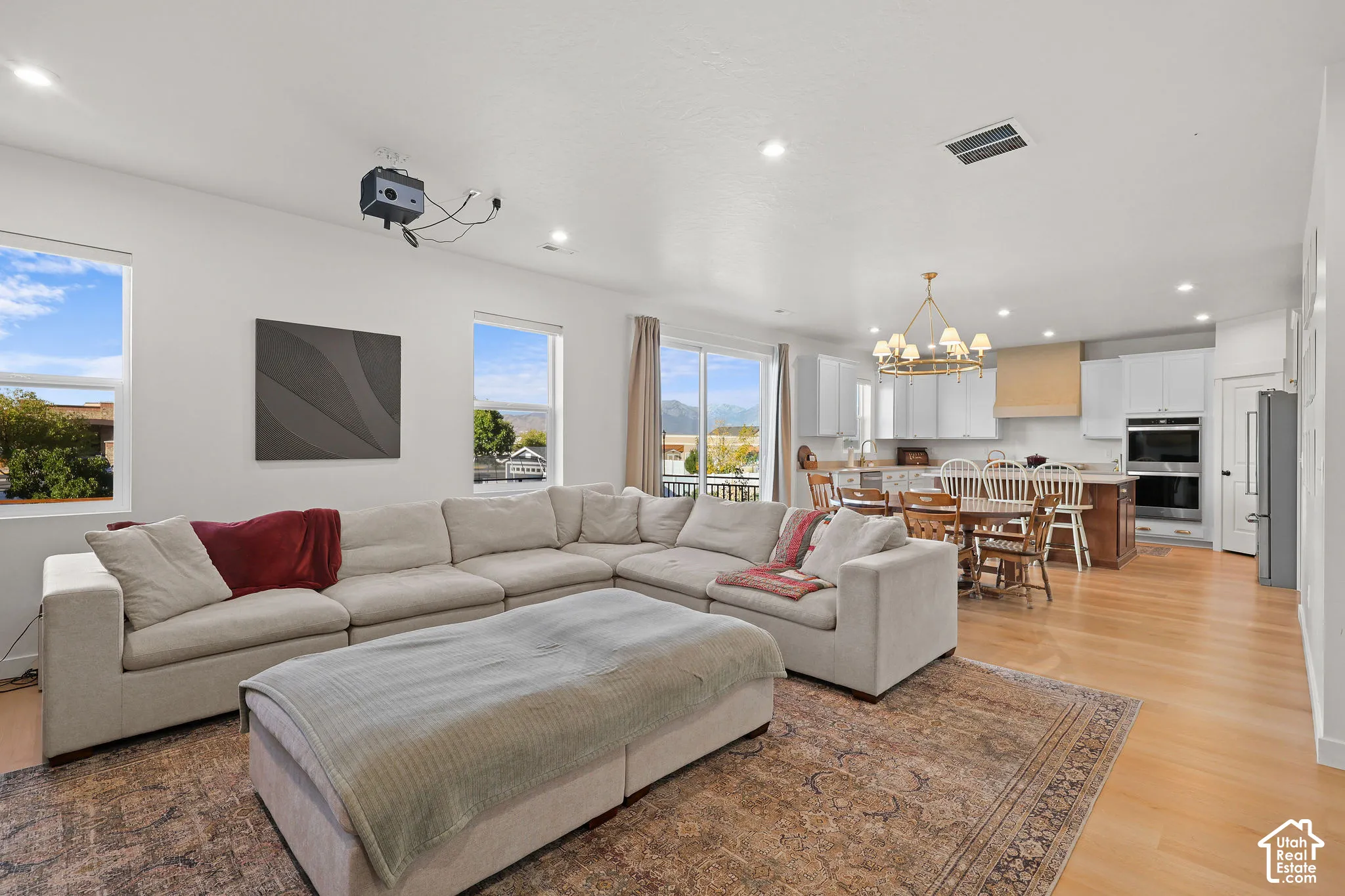 Open-concept interior featuring light wood-finish flooring, recessed lighting, double wall ovens, a custom range hood, and large windows with mountain scenery