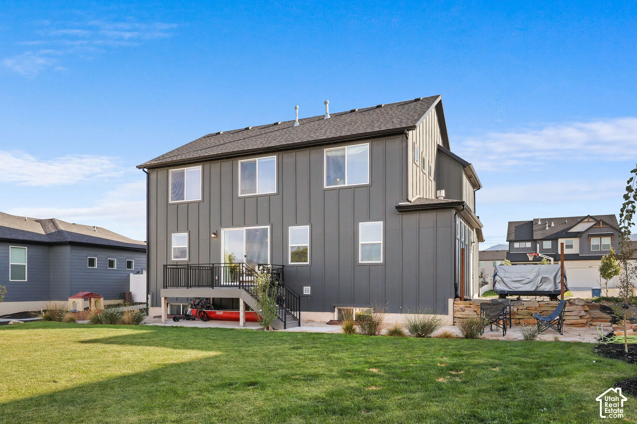 Two-story exterior featuring vertical metal siding, an elevated deck with black metal railings, a concrete patio, and stacked stone retaining wall