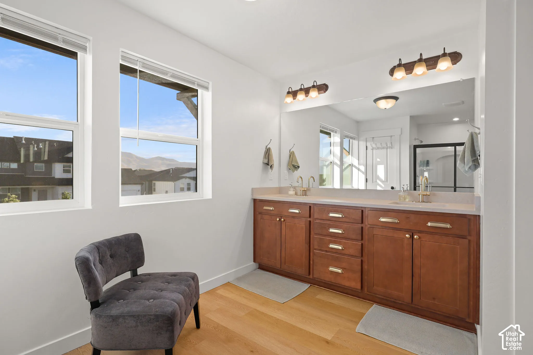 Dual-vanity cabinetry with gold-tone hardware, expansive wall-to-wall mirror, wood-finish flooring, and dual light fixtures featuring glass shades