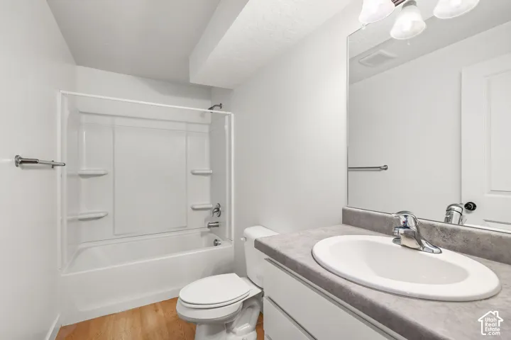 Bathroom featuring a vanity with a solid-surface countertop, an integrated oval sink, a wall-to-wall mirror, a tub-shower combination with built-in shelving, and wood-finish flooring