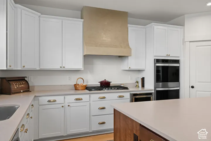 Kitchen featuring white shaker-style cabinetry, a custom plaster-finish range hood, stone-surface countertops, built-in double wall ovens, and a wood-finish central island