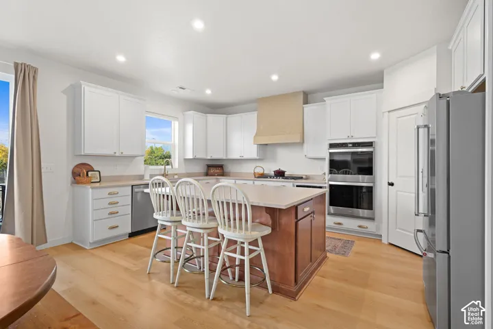 Kitchen featuring white cabinetry, wood-finish flooring, a central island with a contrasting wood base, built-in stainless steel double ovens, and a custom range hood