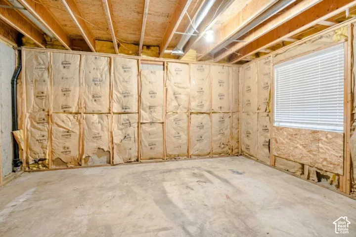 Unfinished room featuring exposed wooden joists, concrete flooring, framed wall studs with batt insulation, and a single window with horizontal blinds
