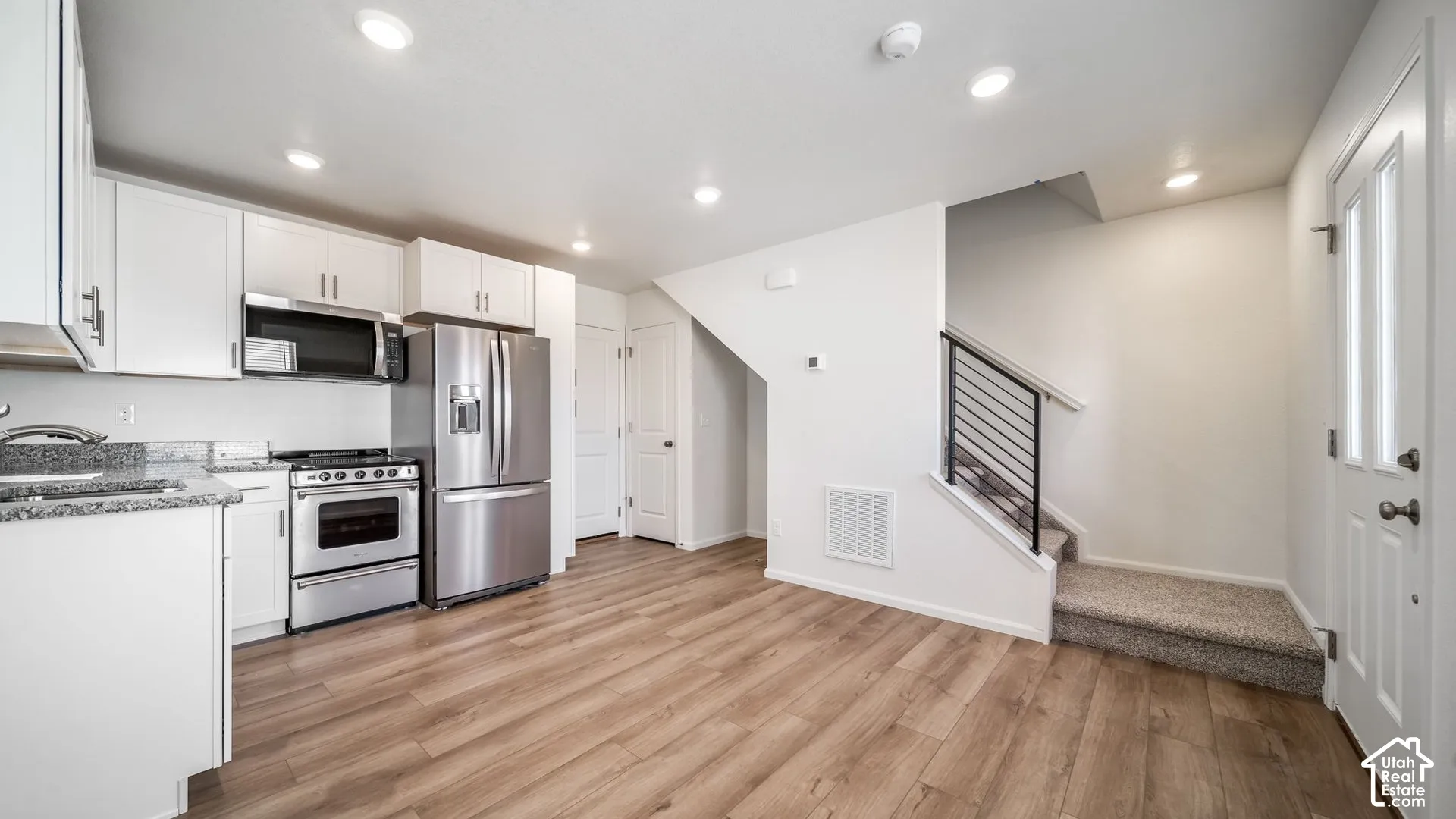 Kitchen featuring white cabinetry, stone-surface countertops, stainless steel appliances, wood-finish flooring, and a staircase with a black metal railing