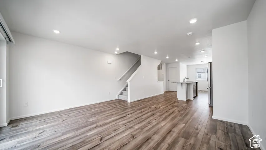 Open-concept interior featuring wood-finish flooring, recessed lighting, a center island with a stainless steel sink, and a staircase with a white handrail