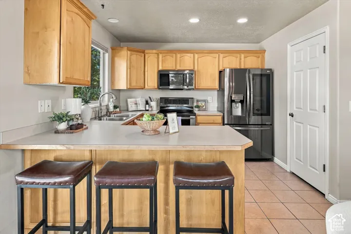 Kitchen featuring natural-finish wood cabinetry, a peninsula with a solid-surface countertop, tile flooring, a stainless steel refrigerator, and a built-in microwave