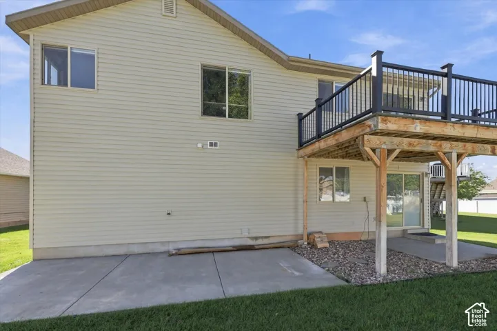 Rear exterior featuring light-colored horizontal siding, an elevated wood deck with black metal railing, a concrete patio, and a grass lawn