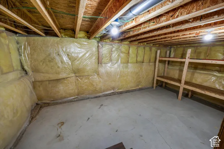 Unfinished storage area featuring exposed wood joists, concrete flooring, and fiberglass wall insulation with a plastic vapor barrier