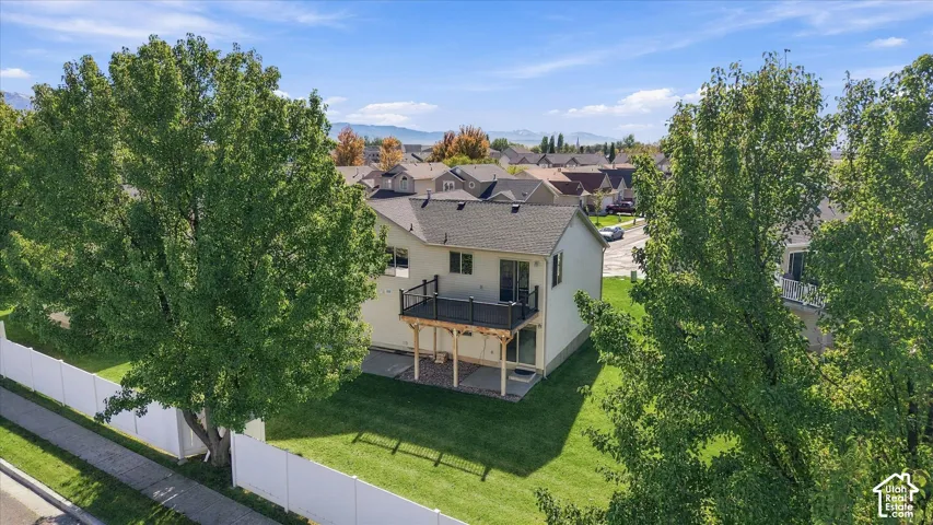 Elevated rear exterior featuring a raised deck with metal railing, concrete ground-level patio, vinyl perimeter fencing, and mature trees