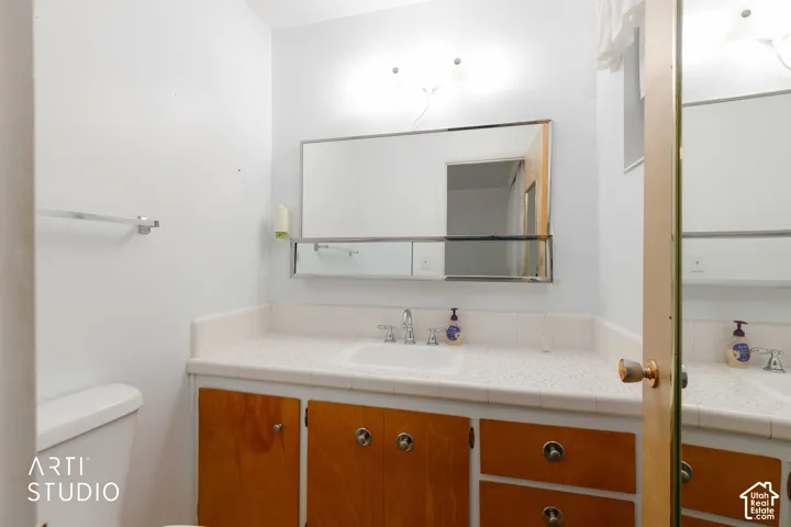 Bathroom vanity featuring wood-finish cabinetry, a white countertop with a tiled backsplash, an integrated rectangular sink, a wide wall-mounted mirror with an integrated shelf, and dual-light fixture