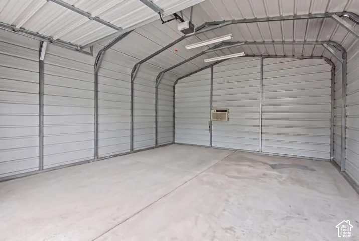 Detached metal garage featuring a concrete slab foundation, high-vaulted ceiling, dual overhead fluorescent light fixtures, and a wall-mounted climate control unit