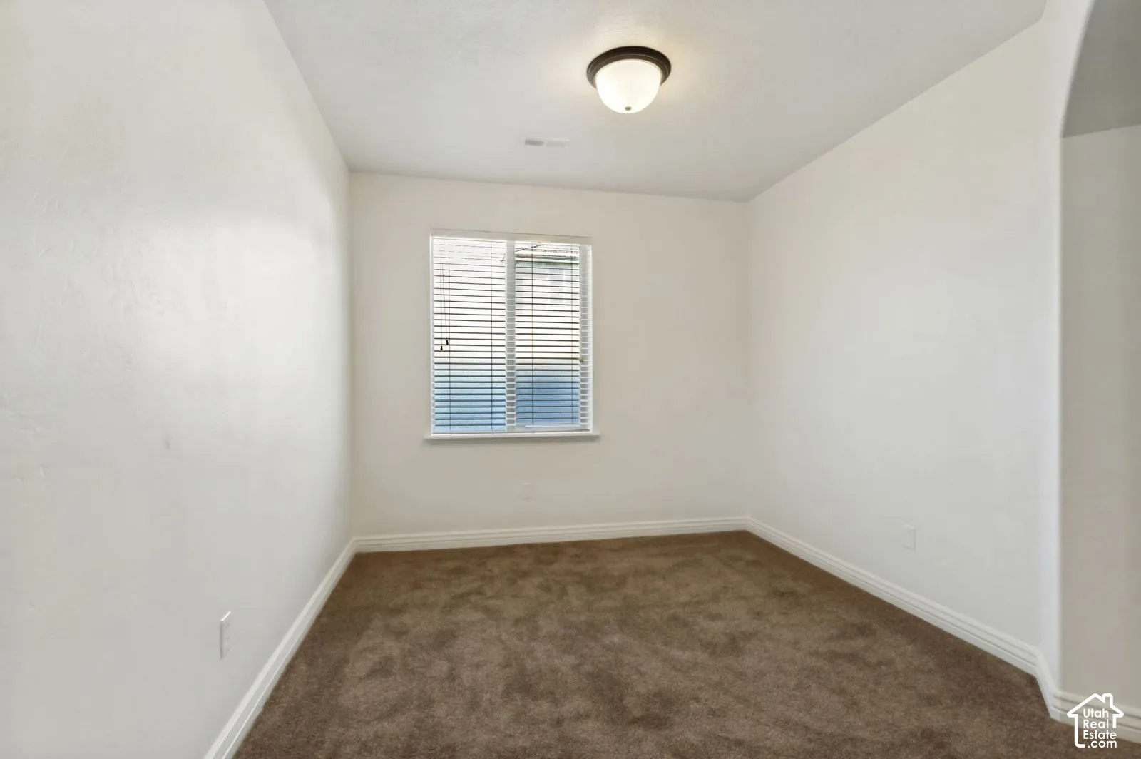 Neutral-toned interior space featuring carpeting, smooth wall finish, single window with horizontal blinds, and a dome-style flush-mount ceiling light