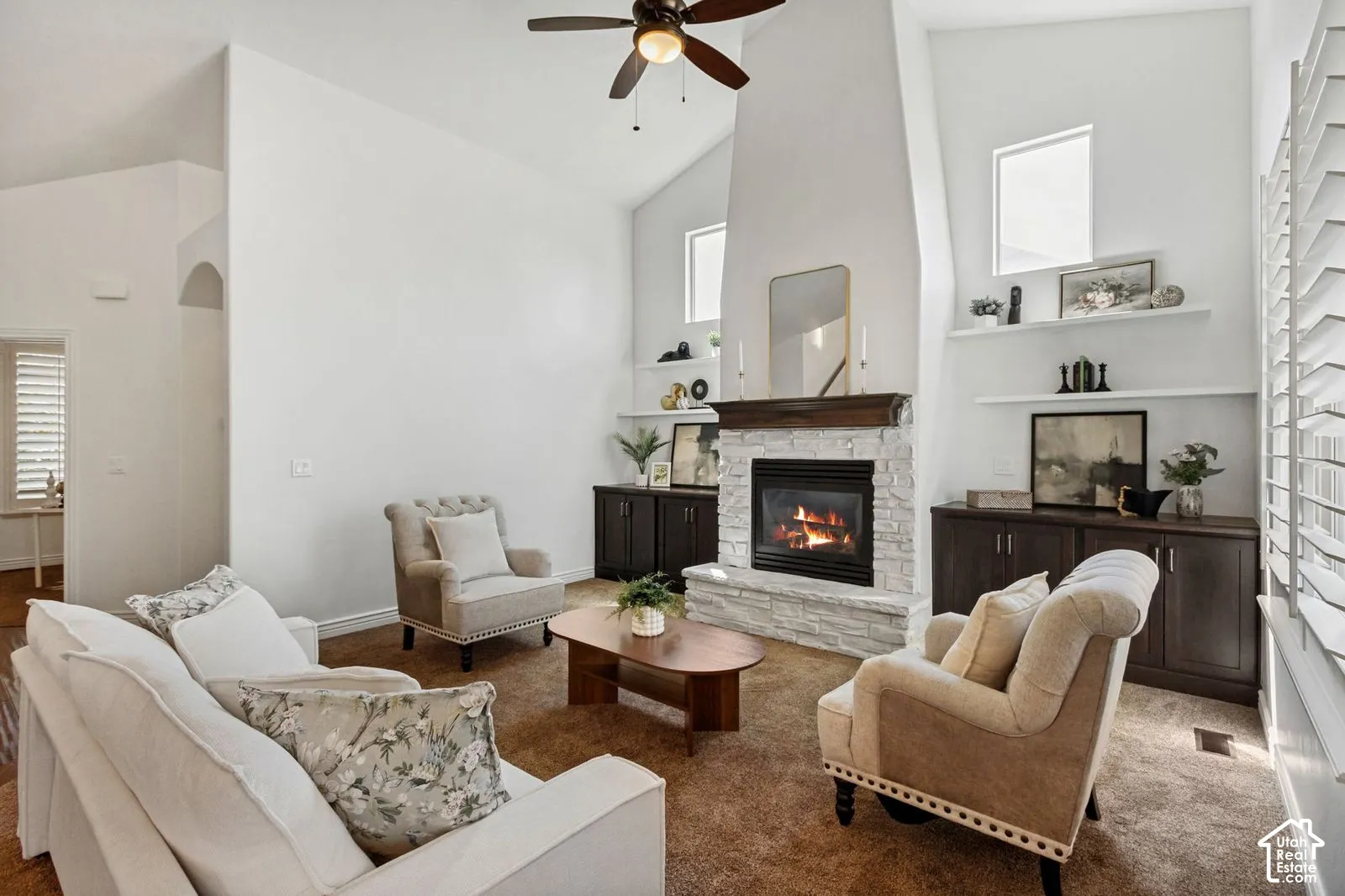 Vaulted ceilings featuring a floor-to-ceiling stone fireplace, dark-finish custom built-in cabinetry, floating display shelving, and an integrated ceiling fan