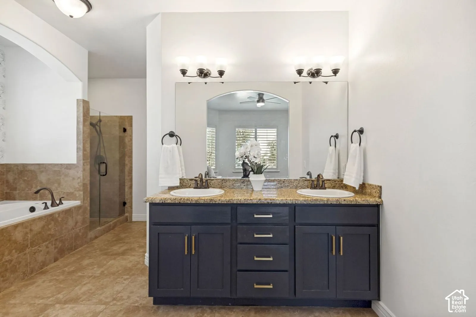 Dual-sink vanity featuring dark-toned cabinetry, light-colored stone-surfaced countertops, and brushed-finish hardware, paired with a glass-enclosed shower and an integrated tile-surround bathtub