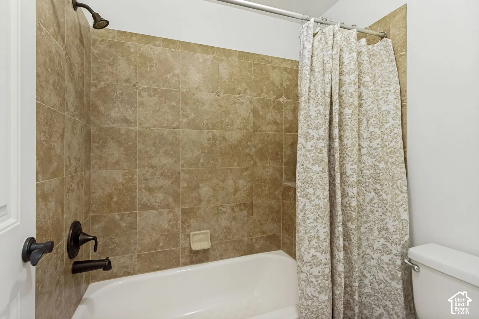 Bathtub surround featuring large-format earth-toned tile, dark-finish wall-mounted faucet and shower head, and built-in soap dish