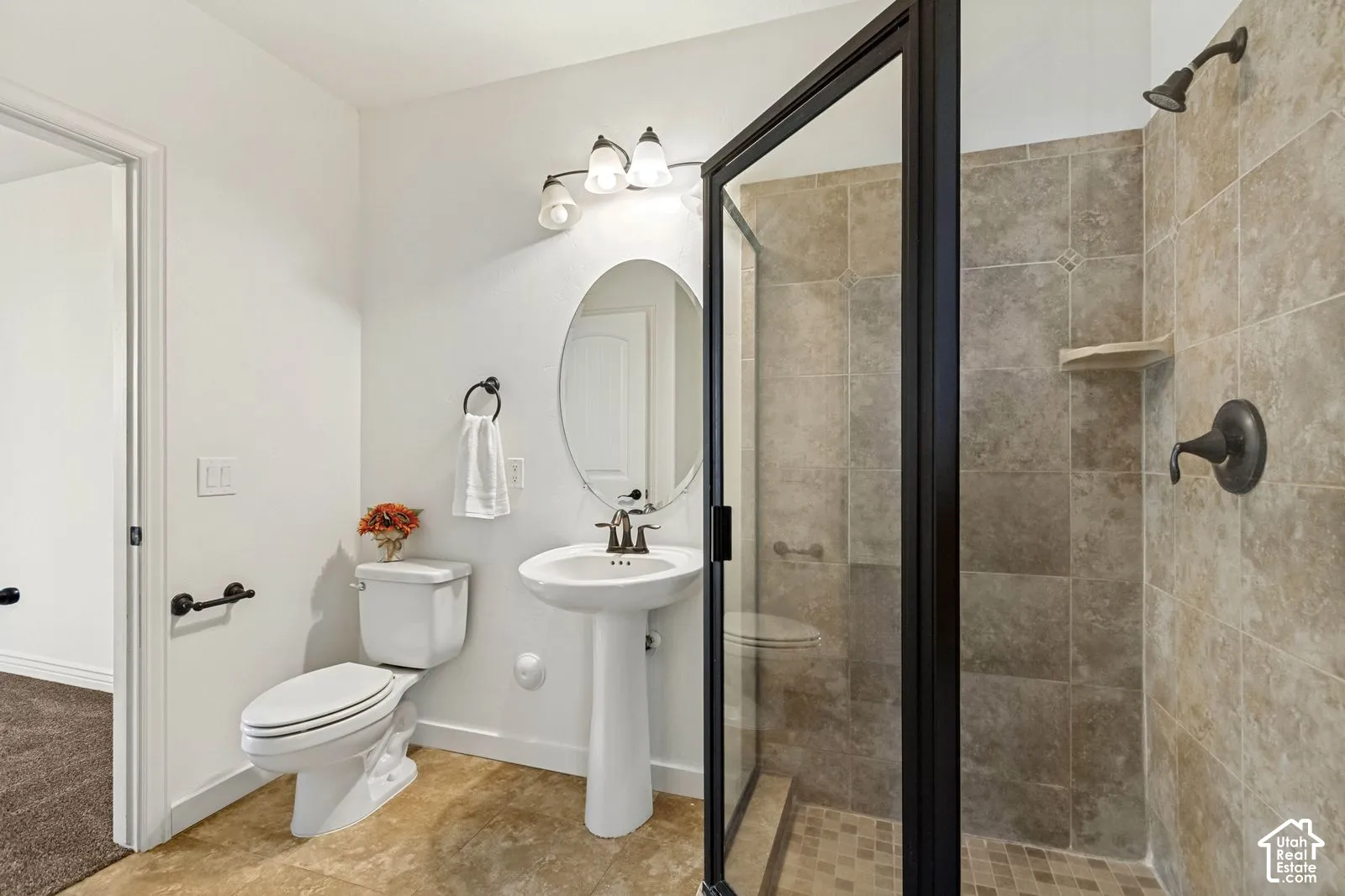 Bathroom featuring a pedestal sink with an oval mirror, three-light vanity fixture, and a tiled shower stall with a black metal-framed glass door and bronze-tone hardware