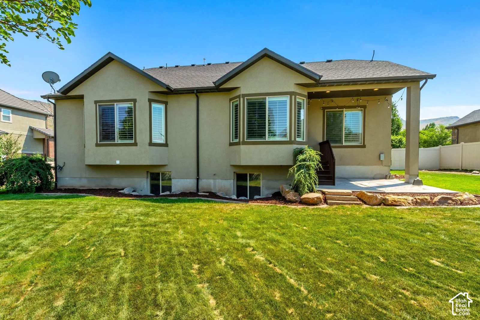 Stucco exterior featuring dual bay windows, a covered concrete patio, tiered stone landscaping, and an expansive lawn