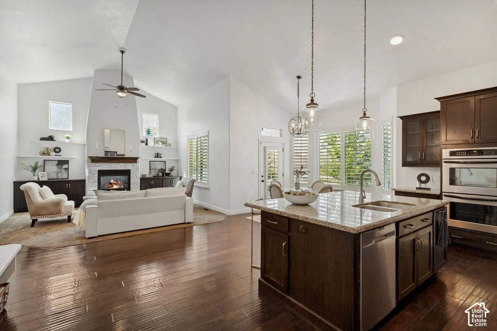 Open-concept living space featuring vaulted ceilings, wood-finish flooring, a kitchen island with stone countertop, stainless steel double wall ovens, and a floor-to-ceiling fireplace with built-in cabinetry