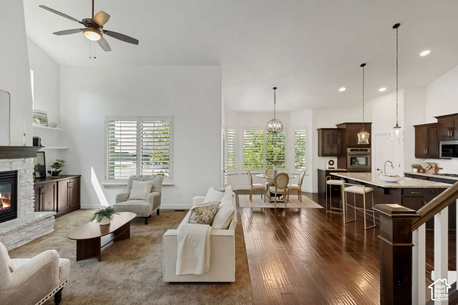 Open-concept floor plan featuring wood-finish flooring, a stacked-stone fireplace with built-in shelving, vaulted ceilings, cabinetry with stone countertops, and plantation shutters