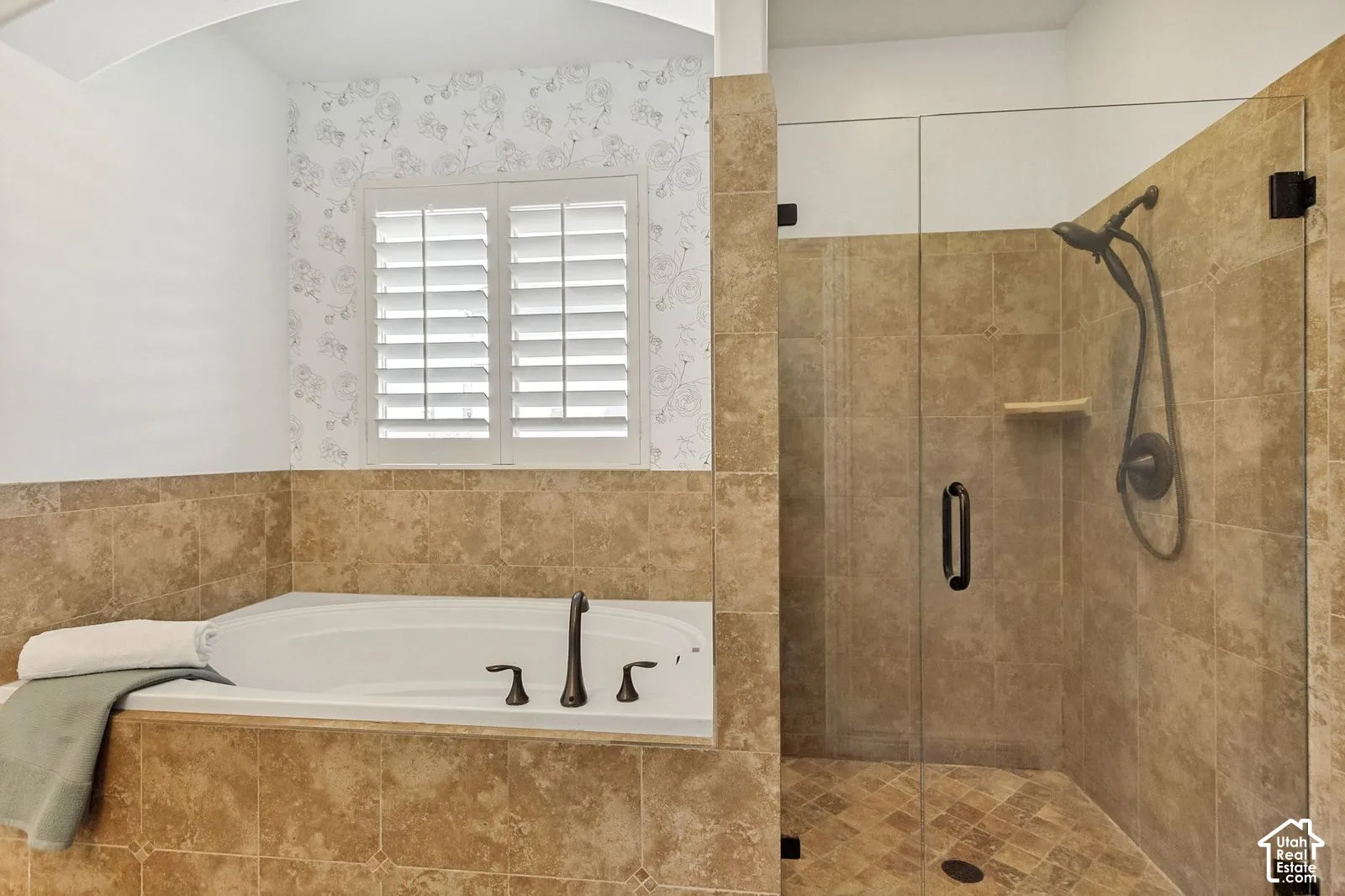 Soaking tub with bronze finish fixtures, walk-in shower with tile surround and glass door, plantation shutters, and patterned wallpaper