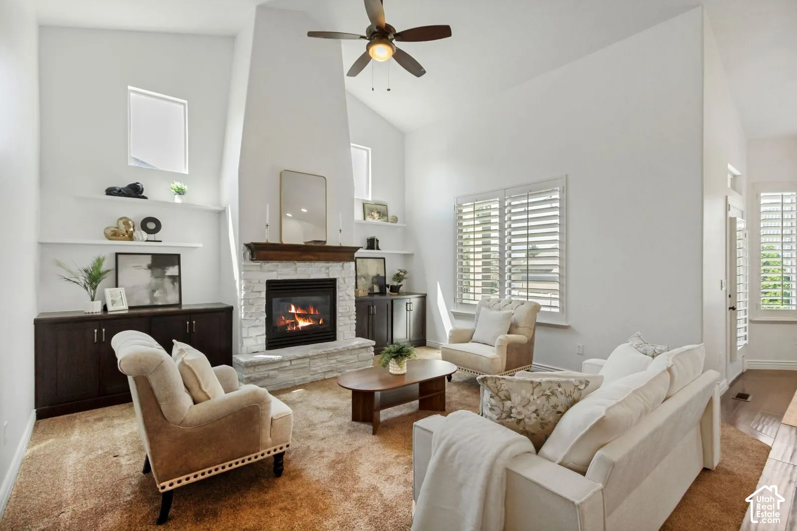 Vaulted ceiling with a central fan, stone-surround fireplace featuring a wood mantel, built-in cabinetry, floating wall shelves, and windows equipped with plantation shutters