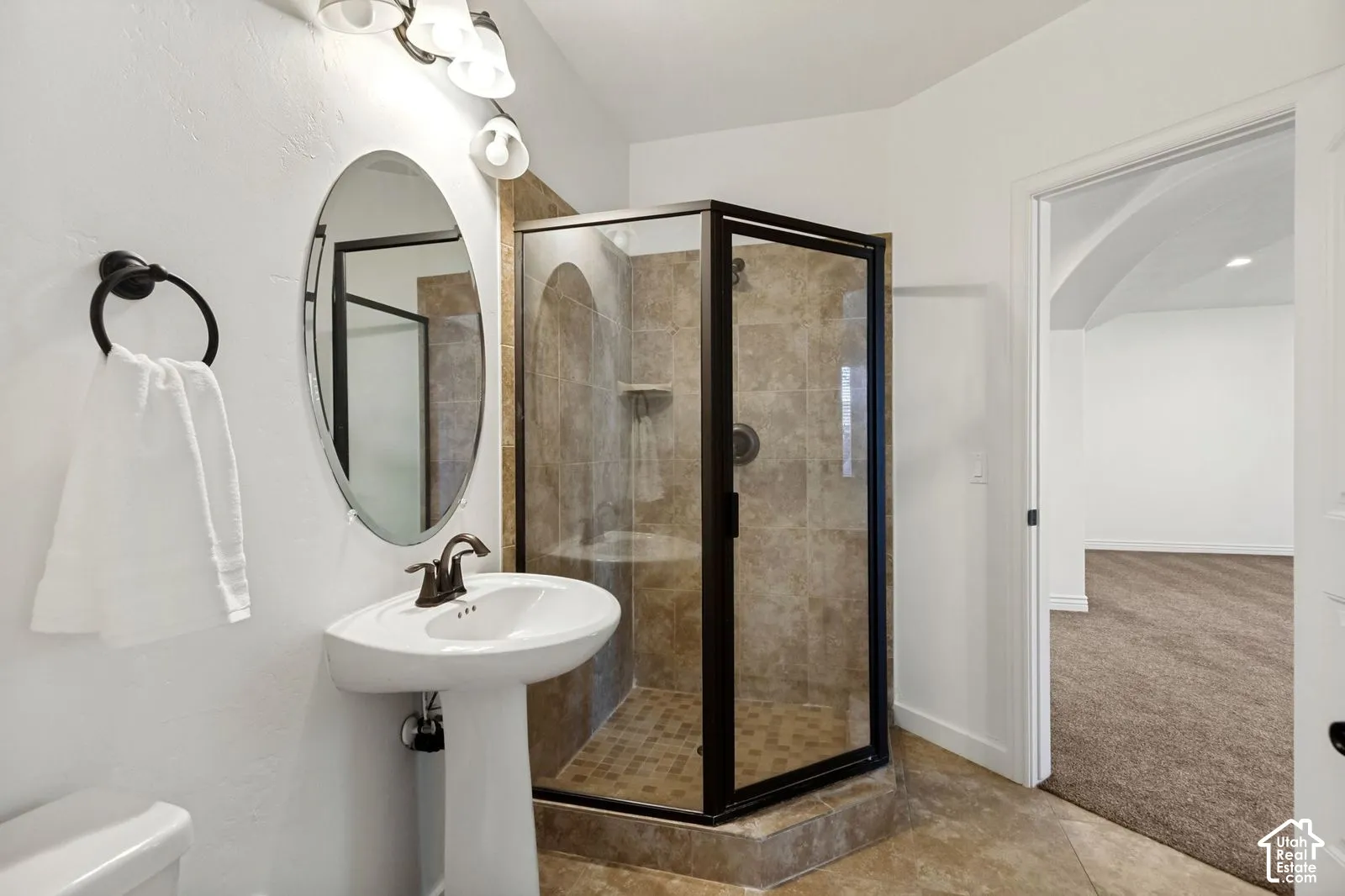 Pedestal sink with a dark metal faucet, oval mirror, and corner shower enclosure with tiled surround